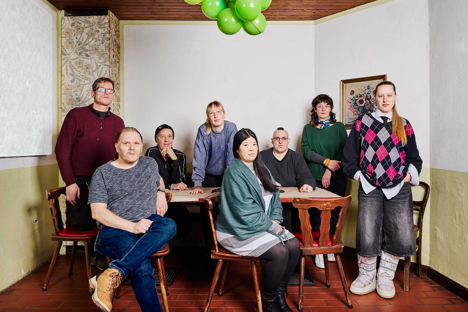 Teamfoto Klappe auf 2025: 8 Personen (4 Frauen und 4 Männer), sitzen um einen Tisch in einem Restaurant. Leicht ernst schauen sie in die Kamera.