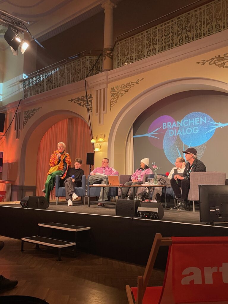 Vier Personen vom Klappe auif Team sitzen im Stuhlkreis auf einer Bühne für einen Vortrag. Festivalleiter der Hofer Filmtage sitzt mit dabei. Auf der Leinwand ist zusehen: Branchen Dialog.