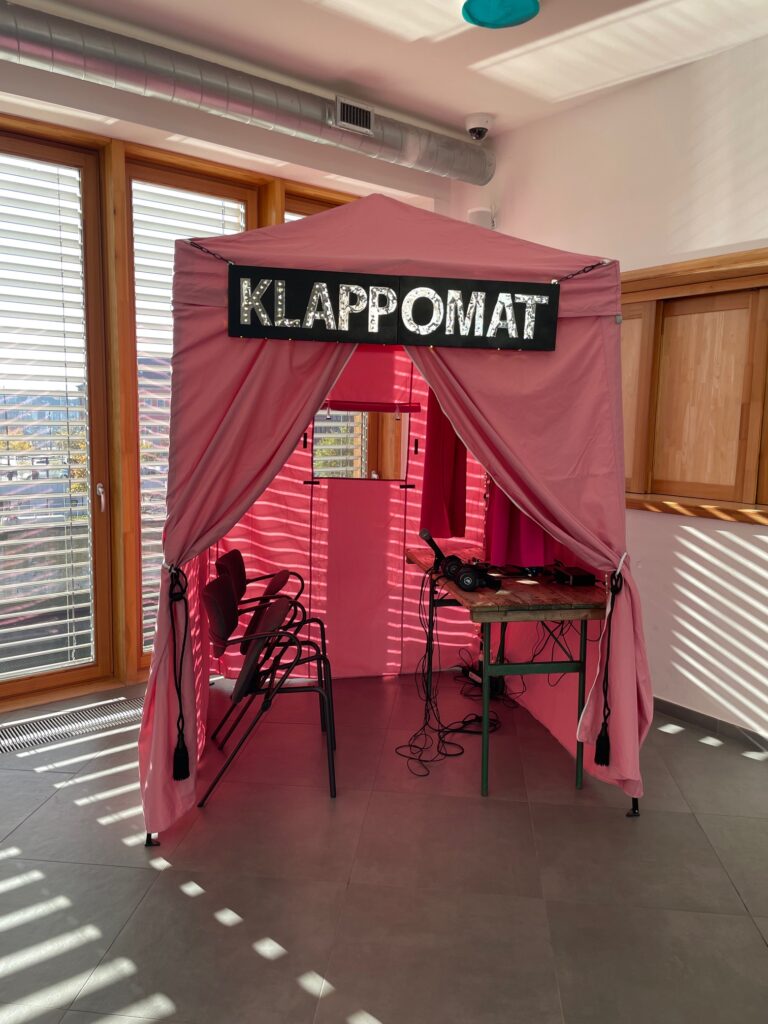 Ein kleiners rosafarbenes Zelt mit der Leuchtschrift Klappomat in einem lichtdurchfluteten Raum. Im Zelt stehen zwei Stühle und ein Tisch mit Technik und Kopfhörern.